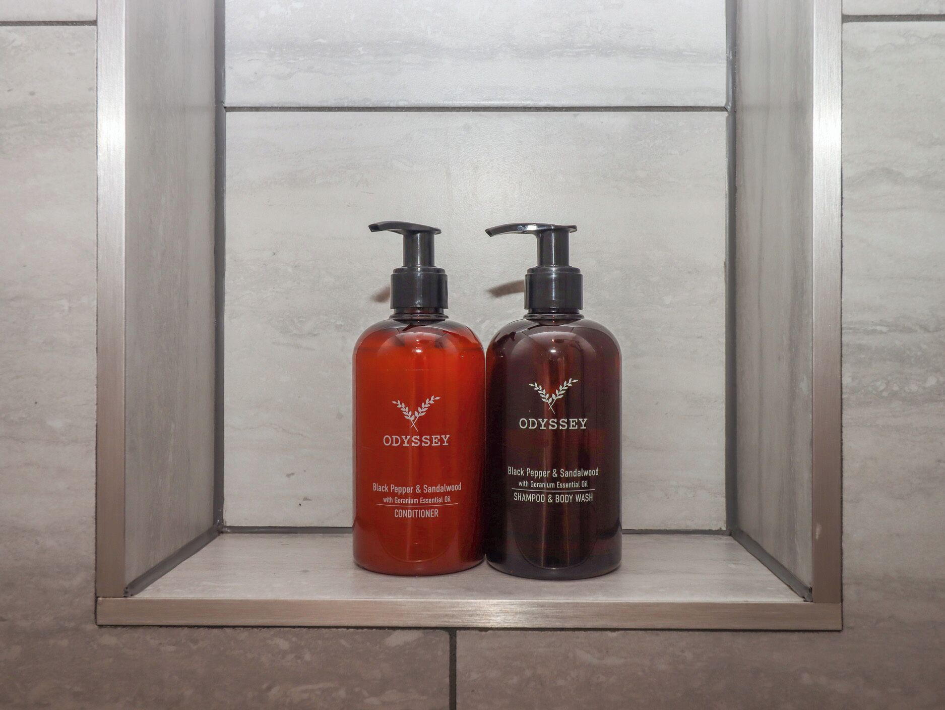 Zwei Flaschen Odyssey Black Pepper & Sandalwood Conditioner und Duschgel auf einem Ablagefach im Badezimmer.