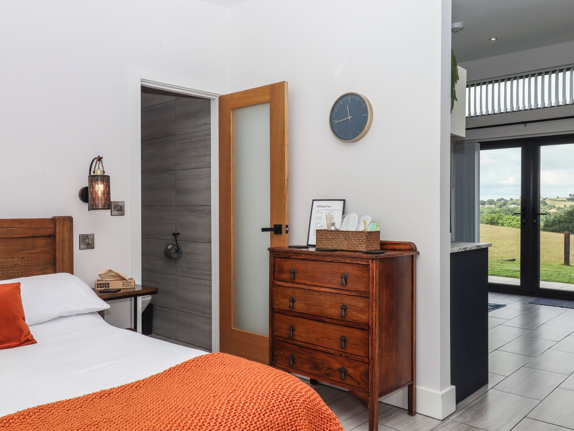 A bedroom with a bed, a wooden dresser, a wall clock, and a door leading to a balcony with a view.