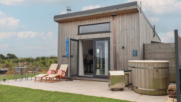 A modern wooden building with a glass door entrance, outdoor seating, and a hot tub. - The Cadeleigh (Tiverton)