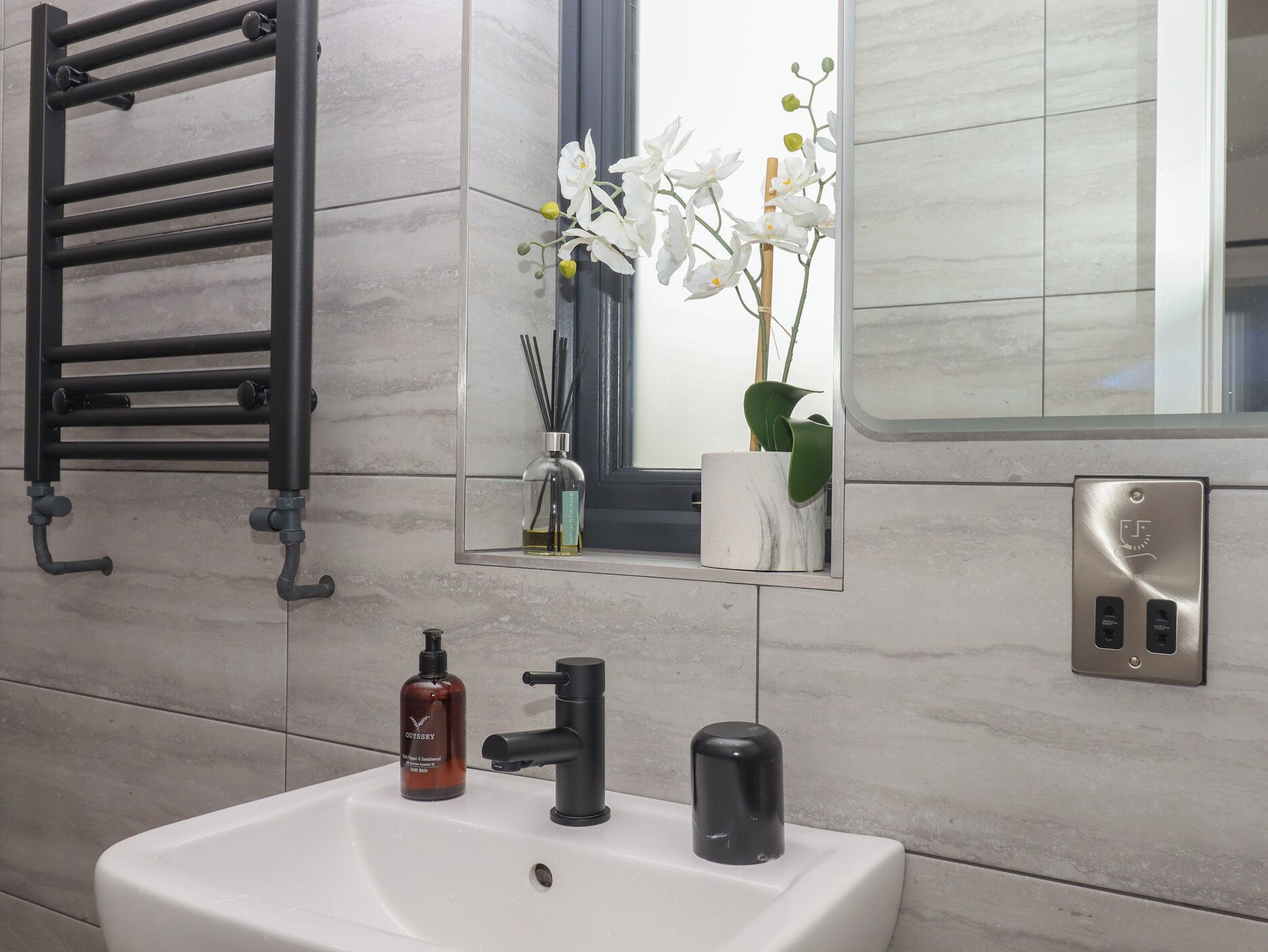 A bathroom with a white sink, black faucet, and a towel rack. There is a mirror with a plant and a bottle of soap.