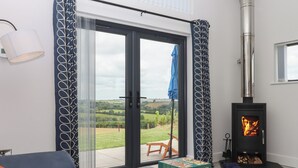 A modern living room with a sofa, a table with a game, a fireplace, and a view of the countryside through large glass doors. - The Cadeleigh (Tiverton)