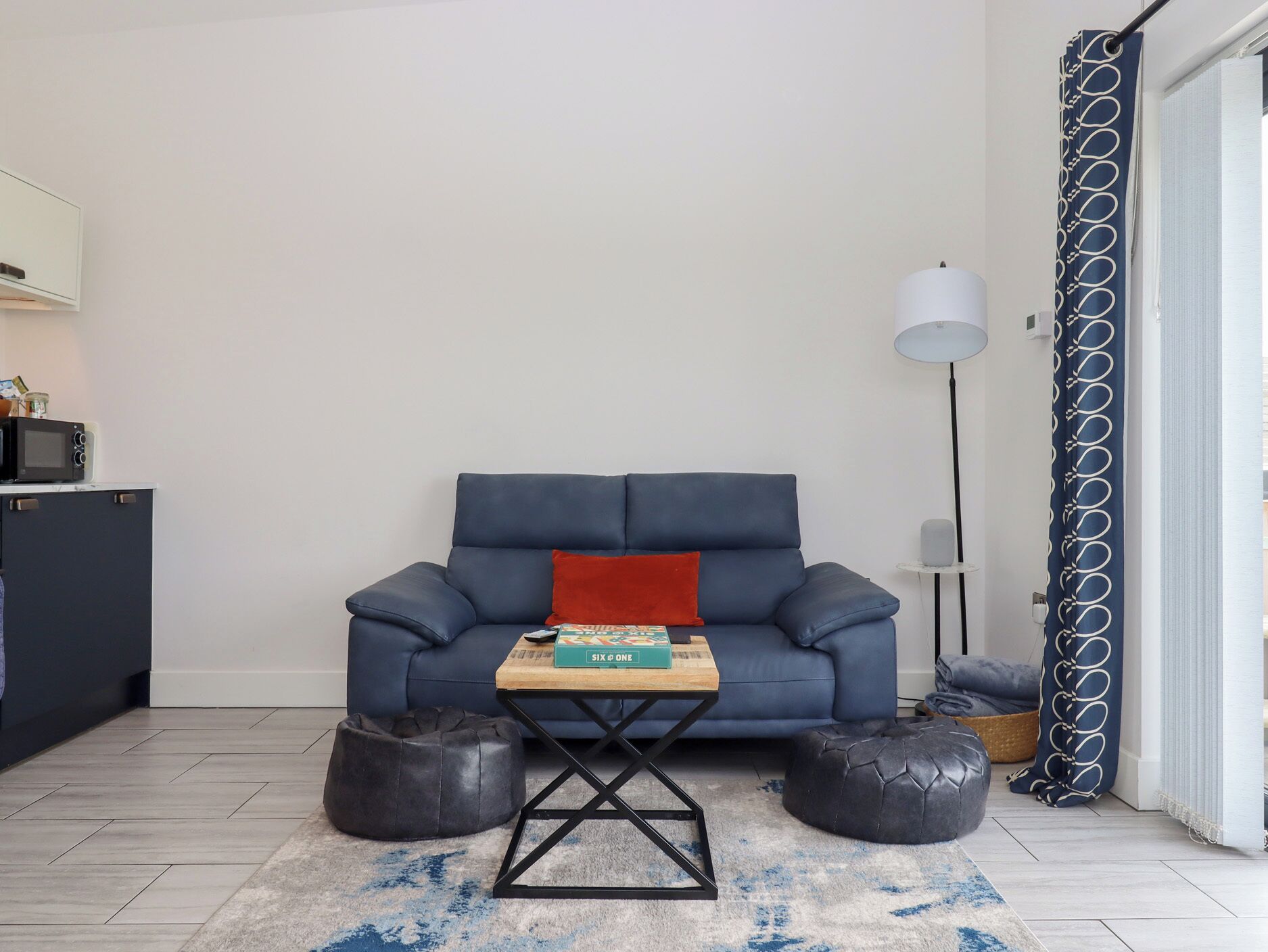 A living room with a blue sofa, a small wooden table with a book, two black ottomans, a grey rug, and a floor lamp.
