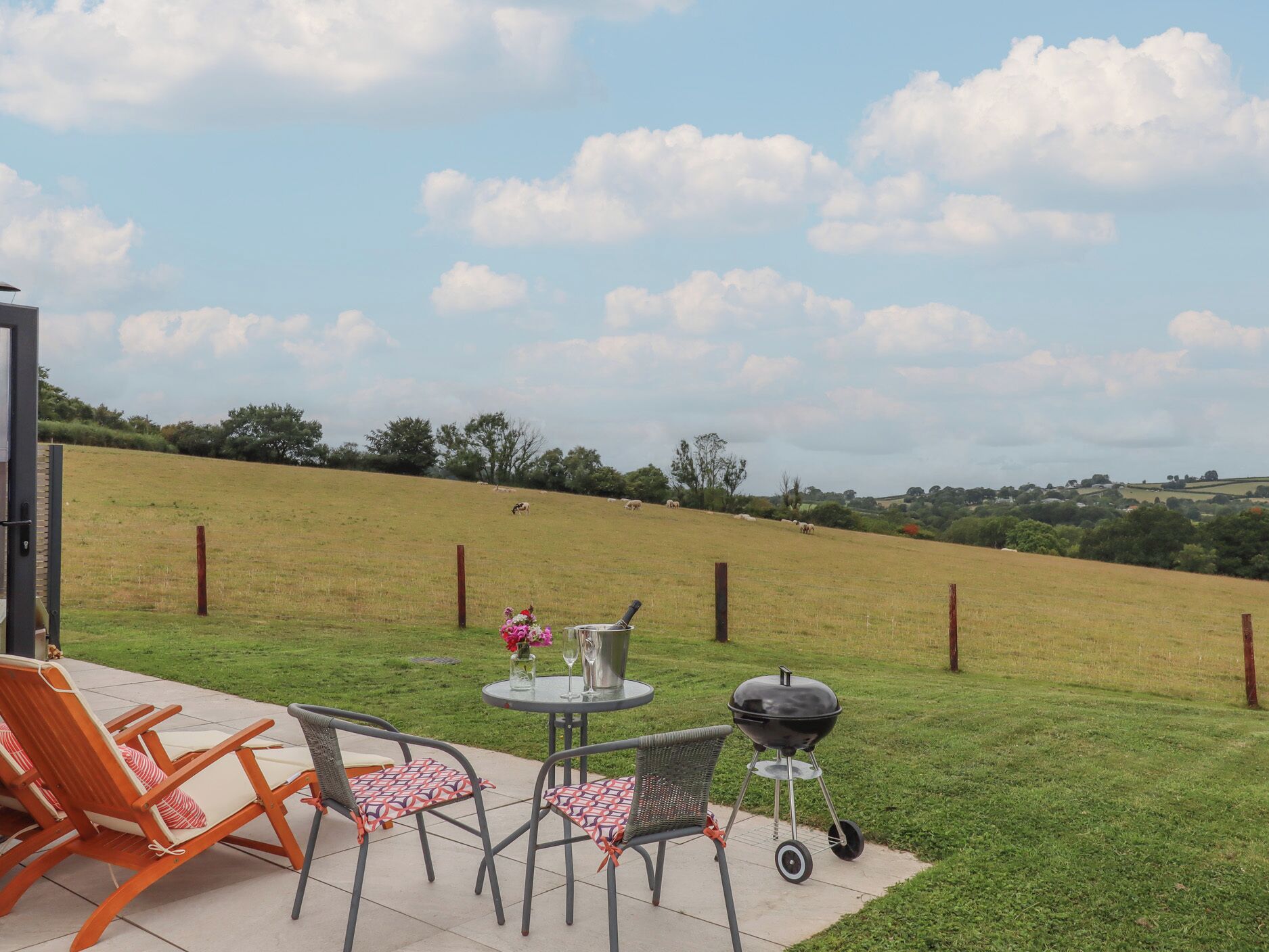 Un patio al aire libre con una mesa, sillas, una parrilla y vistas a un campo cubierto de césped con árboles y colinas al fondo.