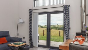A modern living room with a sofa, a coffee table, a lamp, a dining table with chairs, and a view of a grassy field through a glass door. - The Cadeleigh (Tiverton)