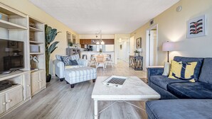 Een ruime woonkamer met een bank, fauteuil en salontafel. Er is een eettafel met stoelen, een keuken en een grote plant.