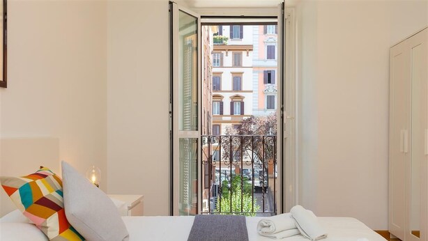 Une chambre d’hôtel avec un lit, des oreillers, une table de chevet avec une lampe, et une vue sur la ville depuis un balcon.