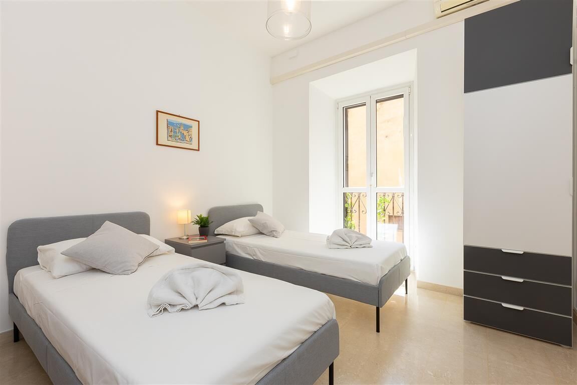 Une chambre avec deux lits, une table de chevet, une commode, une fenêtre et un tableau encadré au mur.