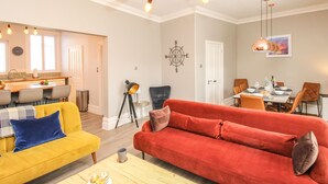 Ein modernes Wohnzimmer mit einem roten Sofa, einem gelben Sessel und einem Holztisch. Im Hintergrund ist ein Essbereich mit Tisch und Stühlen zu sehen.