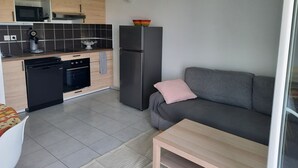 A modern kitchen with a microwave, coffee machine, and dishwasher. There's a gray sofa with a pink pillow and a wooden table. A clock is on the wall. - Bright apartment with large balcony (marseille)