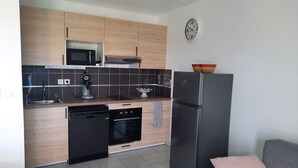 A modern kitchen with wooden cabinets, a microwave, a coffee machine, a dishwasher, an oven, and a refrigerator. There is a clock on the wall and a colorful bowl on top of the refrigerator. - Bright apartment with large balcony (marseille)