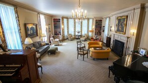 Een ruime woonkamer met een vleugelpiano, comfortabele zithoek, een open haard en een grote boekenkast.