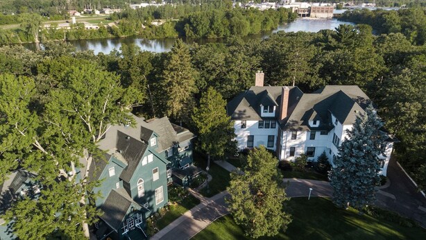Een groot, twee verdiepingen tellend huis met een steile puntige dakkapel, omringd door weelderige begroeiing en een meer.