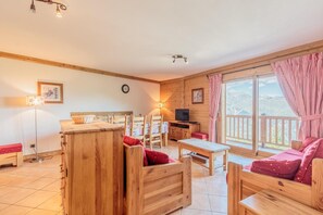 Relax in this inviting room with a large window offering a view of the outdoors, a TV on a wooden stand, and comfortable seating options.
