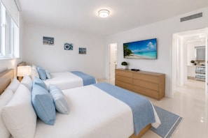 A hotel room with two beds, a wooden dresser, a painting of a beach, and a window with blinds.