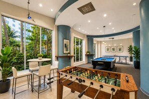 A modern indoor recreational area with a foosball table, pool table, and seating area with a view of greenery outside.