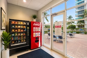 A hotel lobby with vending machines, a Coca-Cola machine, and a view of a pool area with lounge chairs.