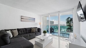 A modern living room with a black sofa, a white coffee table, and a city view through large glass doors. - Wake up over the Atlantic | Private beach access, pool & gym (Hollywood)
