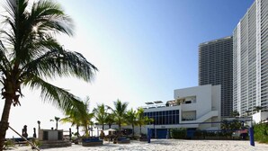 A beach with palm trees, a modern building with large windows, and a tall skyscraper in the background. - Wake up over the Atlantic | Private beach access, pool & gym (Hollywood)