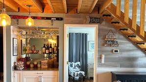 Un interior acogedor con una escalera de madera, una zona de bar con botellas y vasos, y un comedor con una mesa y sillas.