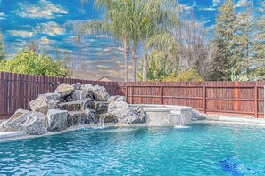 A swimming pool with a rock waterfall feature, surrounded by a wooden fence and trees. - Beautiful Home with Pool & Hot Tub close to National Parks (Selma)