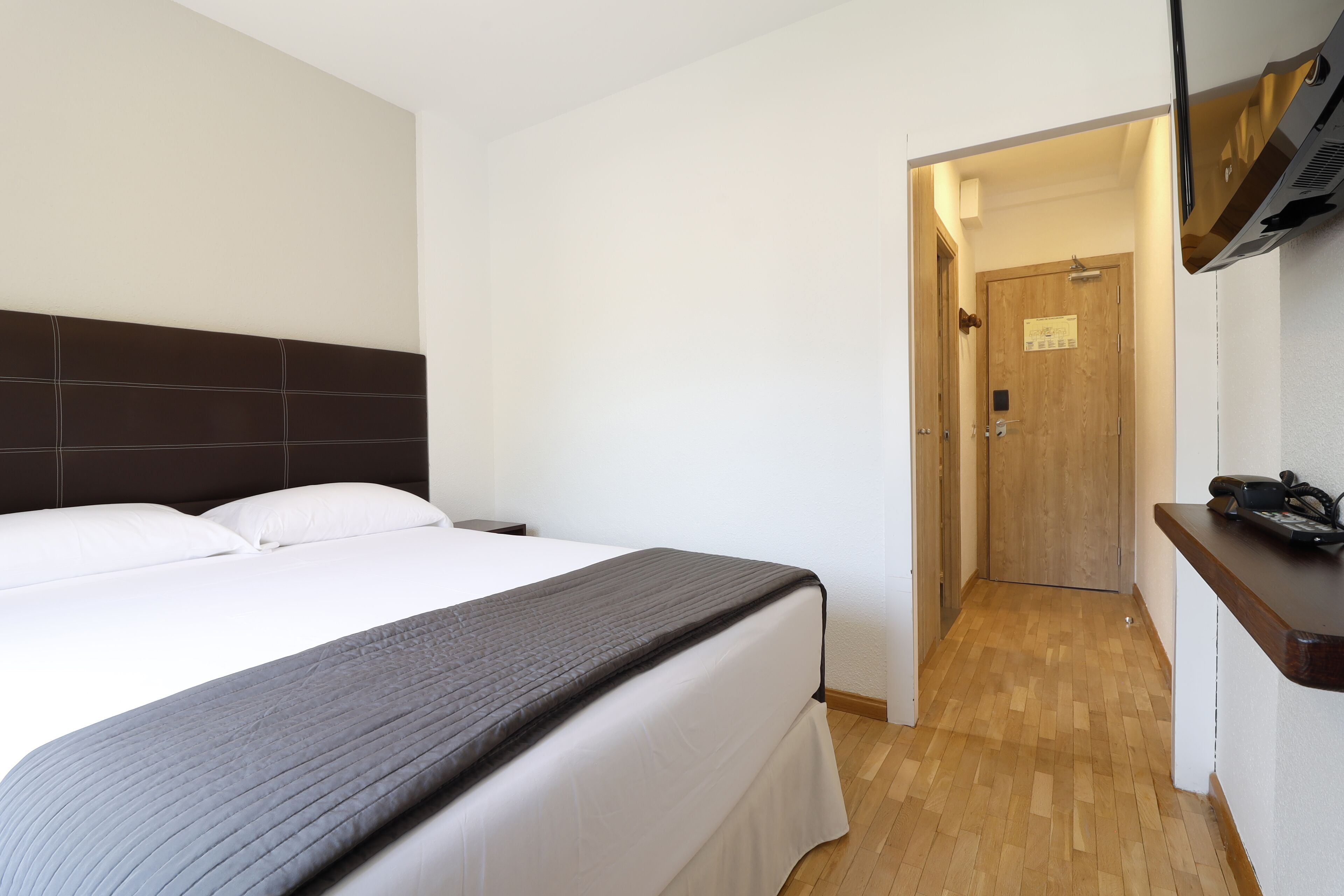 A hotel room with a bed, a TV mounted on the wall, and a wooden floor.