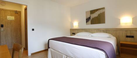 Habitación de hotel con piso de madera, una cama con colcha blanca y morada, cabecera de madera, una lámpara fijada a la pared y una puerta con un letrero.