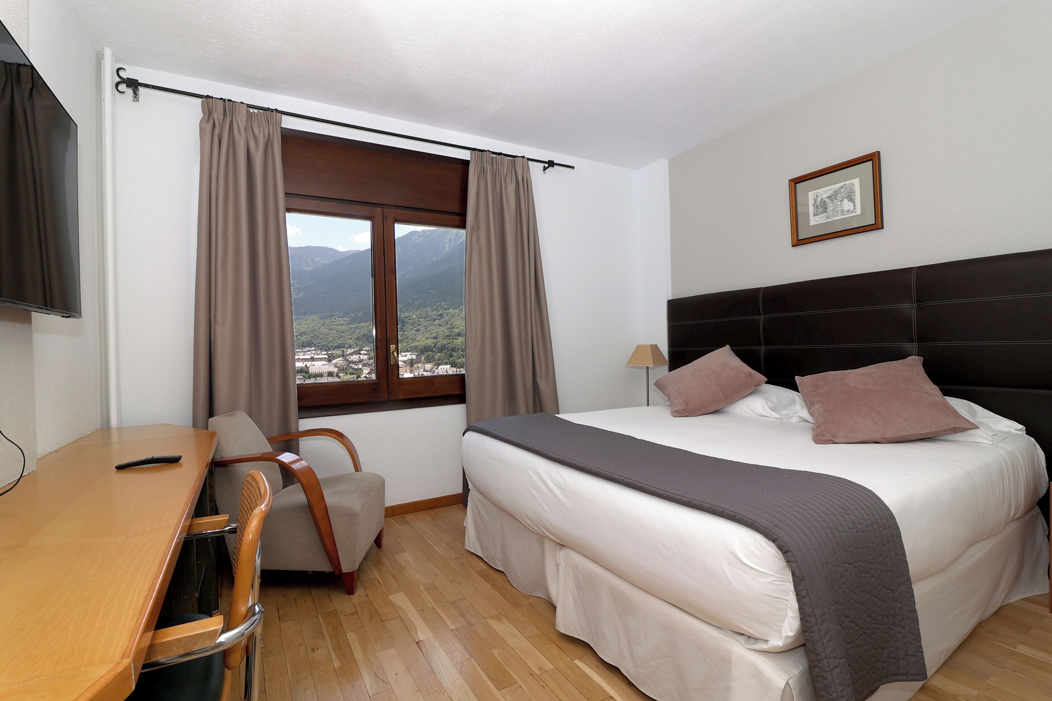 Habitación de hotel con cama, escritorio, silla, ventana con cortinas e imagen enmarcada en la pared.