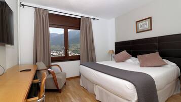 A hotel room with a bed, a desk, a chair, a window with curtains, and a framed picture on the wall.
