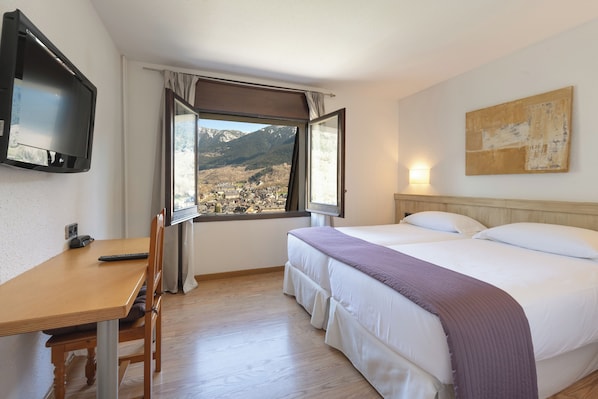 A hotel room with a bed, a desk, a chair, a TV, a window with a mountain view, and a painting on the wall. - Hotel Blu Aran (Vielha e Mijaran)