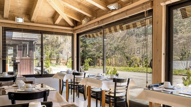 Un comedor con mesas dispuestas para dos, ventanas amplias que ofrecen vistas al exterior y vigas de madera en el techo.