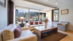 Una sala moderna con sofá, sillón y mesa de centro. Amplias ventanas ofrecen vista a montañas y un estacionamiento.