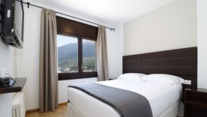 Habitación de hotel con cama, televisor montado en la pared, ventana con cortinas y vista a las montañas.