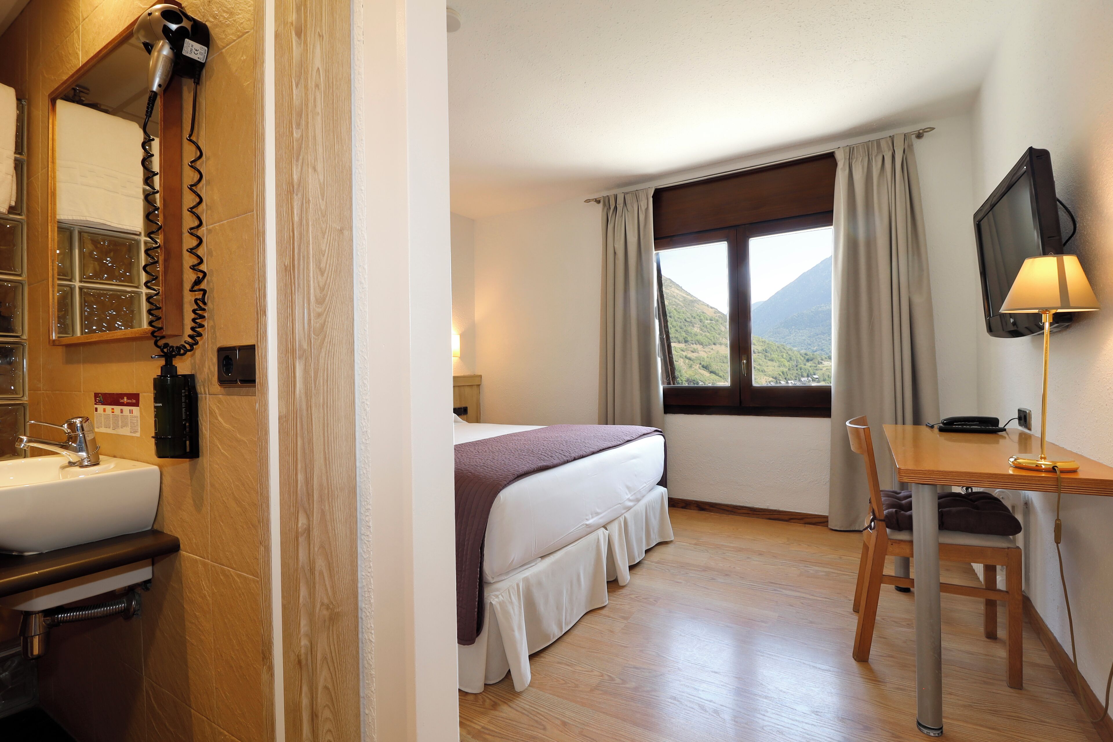 A hotel room with a bed, a desk, a chair, a TV, a lamp, a mirror, and a bathroom with a sink and a hairdryer.