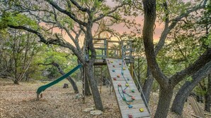 Un'area giochi in legno con una parete da arrampicata e uno scivolo, circondata da alberi.
