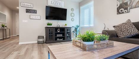 Una sala de estar moderna con un televisor de pantalla plana, una mesa de centro con plantas, un sofá gris y decoración en la pared.