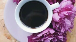 A white cup of coffee on a white saucer surrounded by purple flowers.
