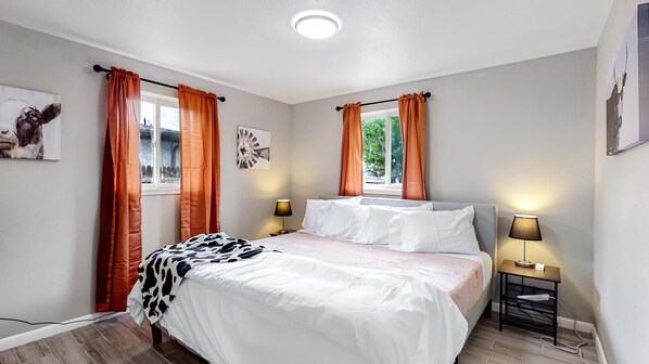 A bedroom with a bed, two bedside tables with lamps, a window with curtains, and a wall clock.