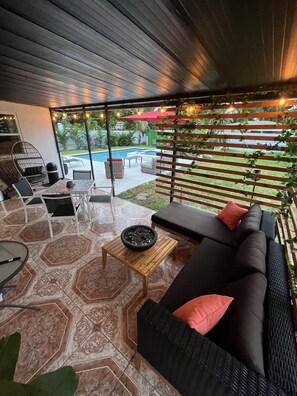 An outdoor patio area with a black sofa, a round table with a bowl, and a wicker chair. - Modern Vacation Home with pool- Conveniently located to Restaurants & Beach (North Miami)