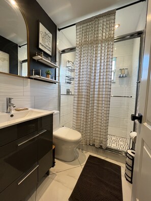 A modern bathroom with a white sink, dark cabinetry, a toilet, a shower with a patterned curtain, and a small shelf with toiletries. - Modern Vacation Home with pool- Conveniently located to Restaurants & Beach (North Miami)