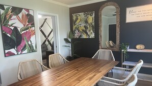 Un coin repas avec une table en bois et des chaises en osier, entouré de grandes œuvres d’art aux motifs tropicaux, d’un miroir et d’une horloge posée sur une étagère.