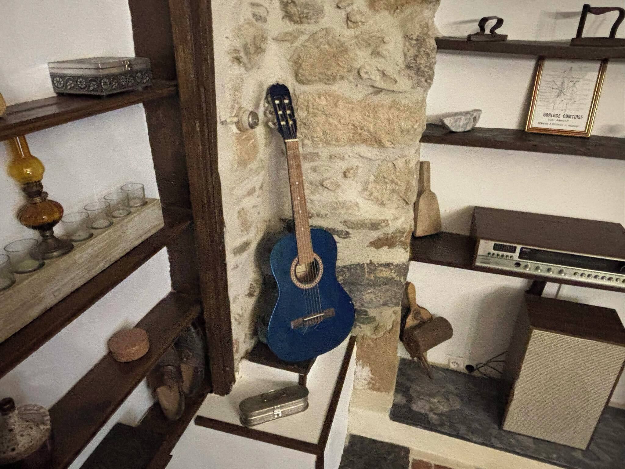 Et værelse med en stenvæg, en blå guitar lænet op ad den, en vintage-radio og et lille bord med en glaskrukke og et stearinlys.