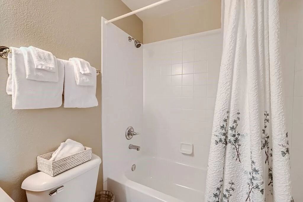 Un baño con inodoro, ducha con cortina blanca y un cesto de artículos de aseo en el inodoro.