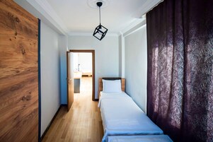 Un pasillo estrecho que conduce a una cama con cabecera de madera y ropa de cama blanca. Una lámpara colgante moderna ilumina el techo.