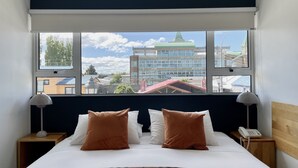 Une chambre d’hôtel avec un lit, deux tables de chevet, un téléphone et une vue sur les immeubles à travers de grandes fenêtres.