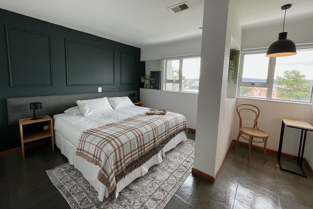 A modern bedroom with a large bed, a side table, a chair, a desk, and a window with a view.