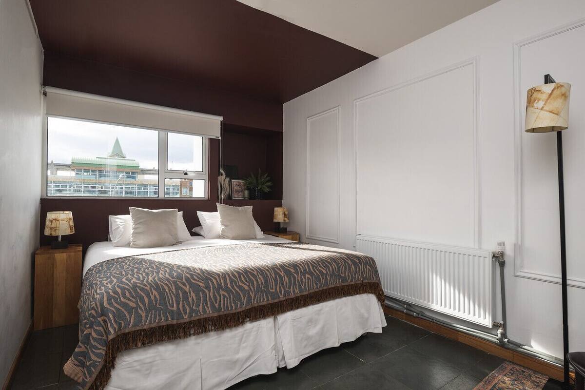 A bedroom with a large bed, a window with a view of a building, a radiator, and a floor lamp.
