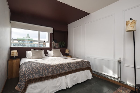 Un dormitorio con una cama grande, una ventana con vistas a un edificio, un radiador y una lámpara de pie.
