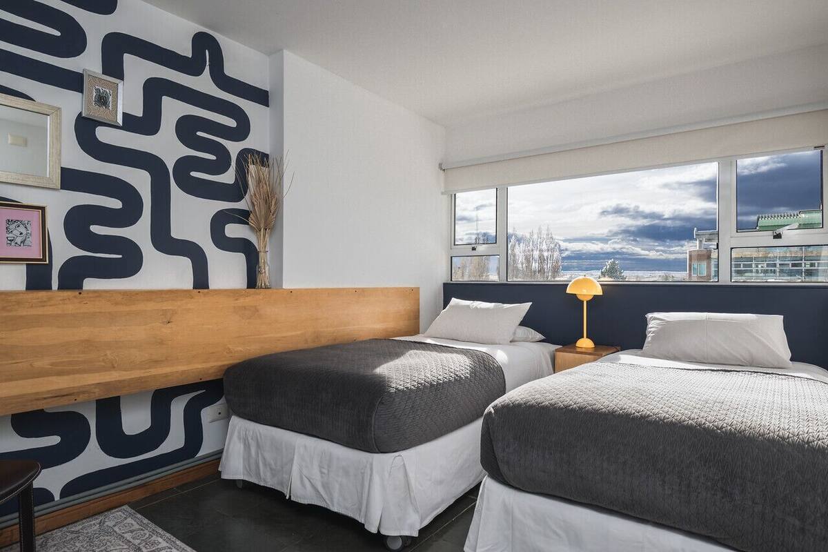 A modern hotel room with a large window, a wooden bench, two beds, and a black and white patterned wall.