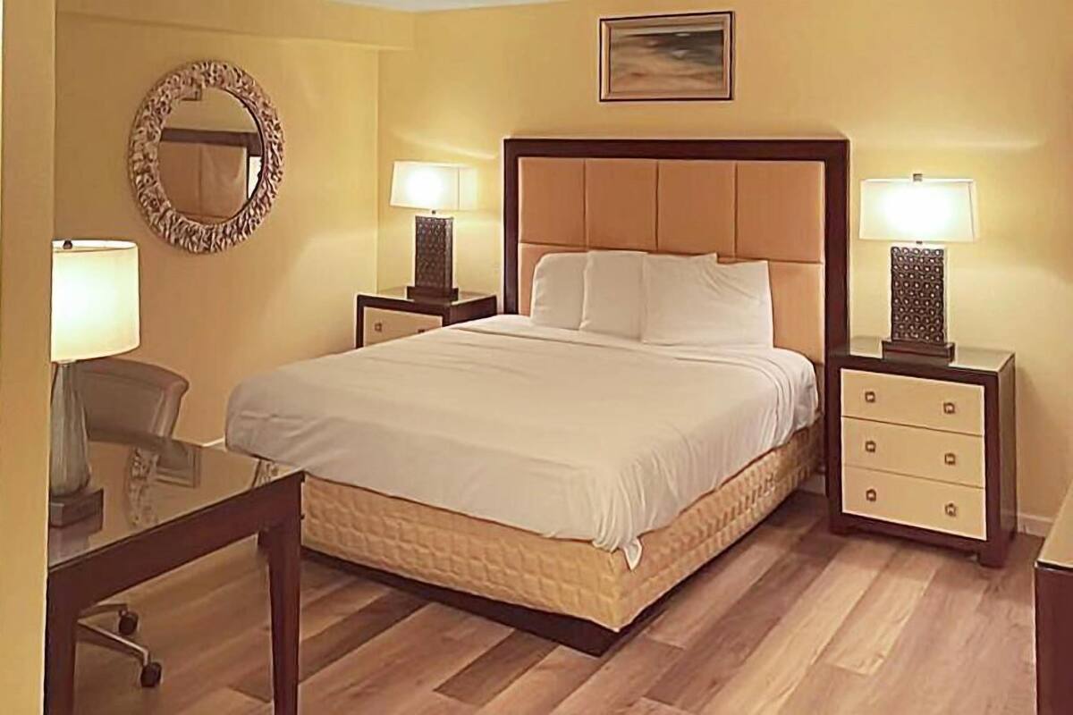A hotel room with a large bed, two bedside tables with lamps, a round mirror, and a small desk with a chair.