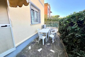 Un pequeño patio exterior con una mesa y sillas, una ventana con rejas y un seto.